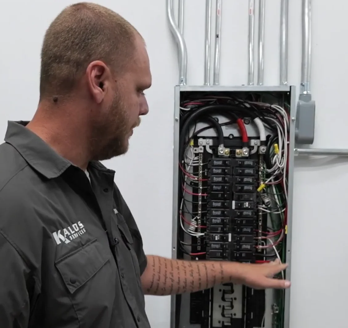 KALOS employee beside an open electrical panel with rows of circuit breakers visible.