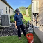 A KALOS plumbing technician beside an exterior tankless water heater, holding service hoses over a bucket during maintenance.