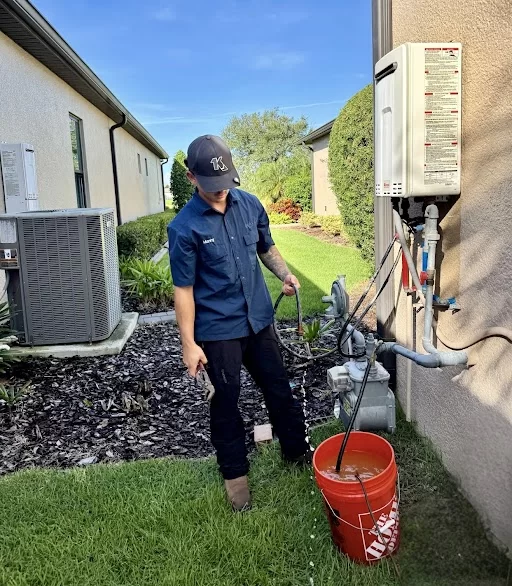 A KALOS plumbing technician beside an exterior tankless water heater, holding service hoses over a bucket during maintenance.